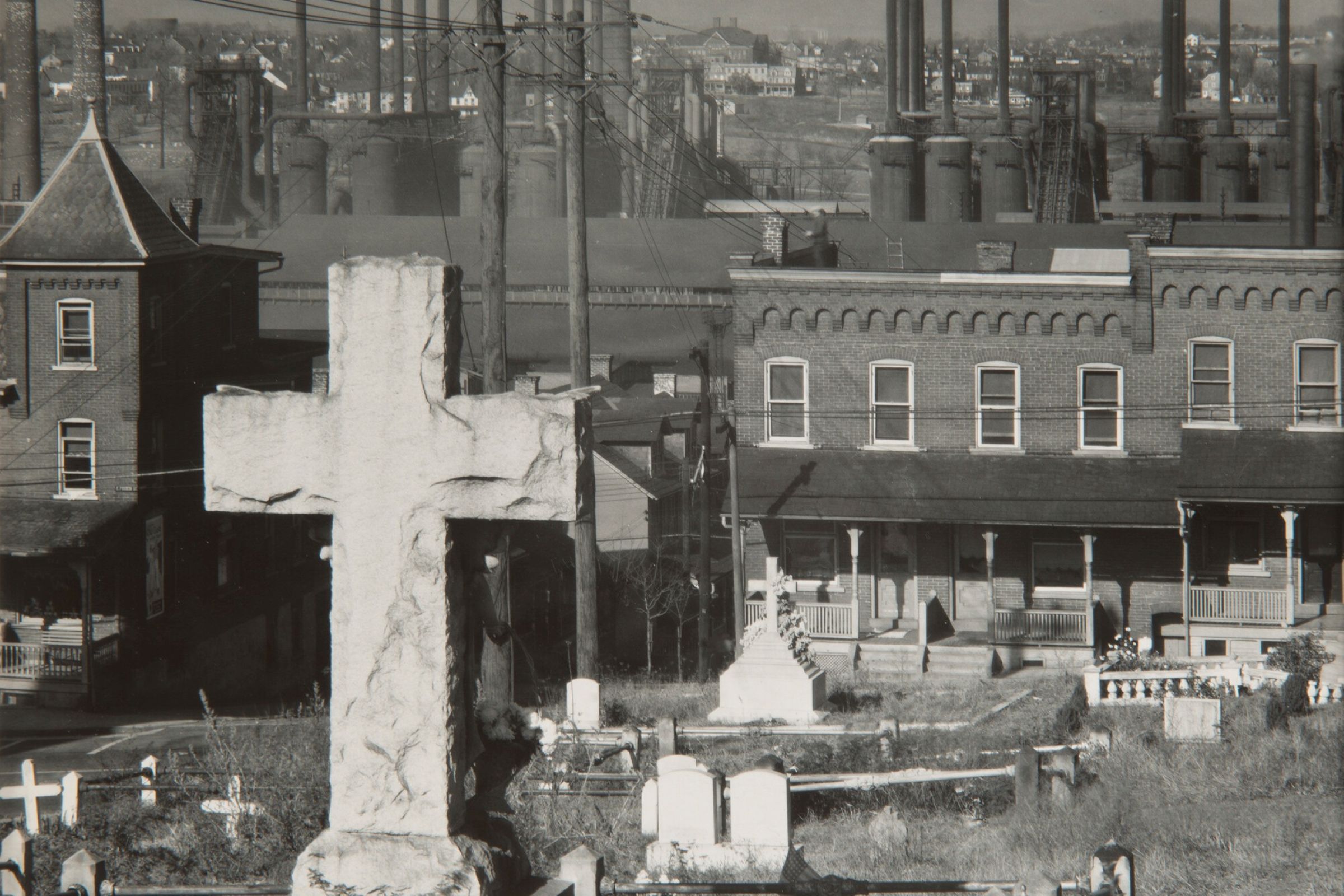 Black-and-white photograph of a cemetery with brick rowhouses and steel mills in the background