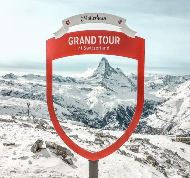 A red, shield-shaped sign stands in the foreground of a snowy mountain landscape. The sign is part of the 'Grand Tour of Switzerland' and features the famous Matterhorn peak in the background. 
