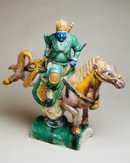 A colorful, detailed statue of a rider on a horse. The figure is dressed in a green and blue outfit, with intricate patterns, and has a blue face and hands. The rider is wearing a crown-like headpiece. The horse, poised in a dynamic stance, has a brown body with a prominent, flowing yellow mane and tail.