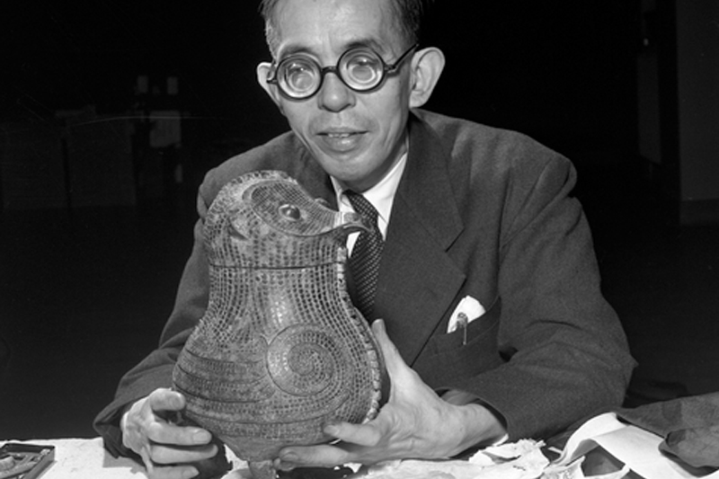 Black-and-white photograph of a person wearing round glasses and a suit jacket seated and holding a decorative ceramic vessel shaped like an owl.