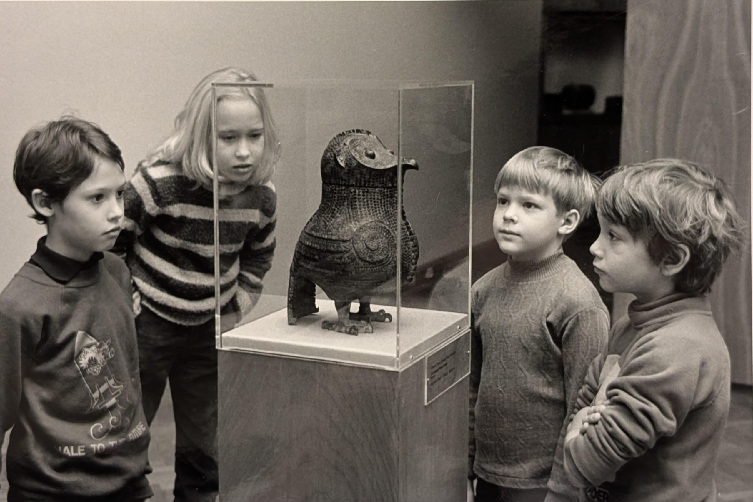 Black-and-white photo of four young children standing closely around a museum display. They are observing an owl-shaped sculpture encased in a transparent glass box set on a pedestal.