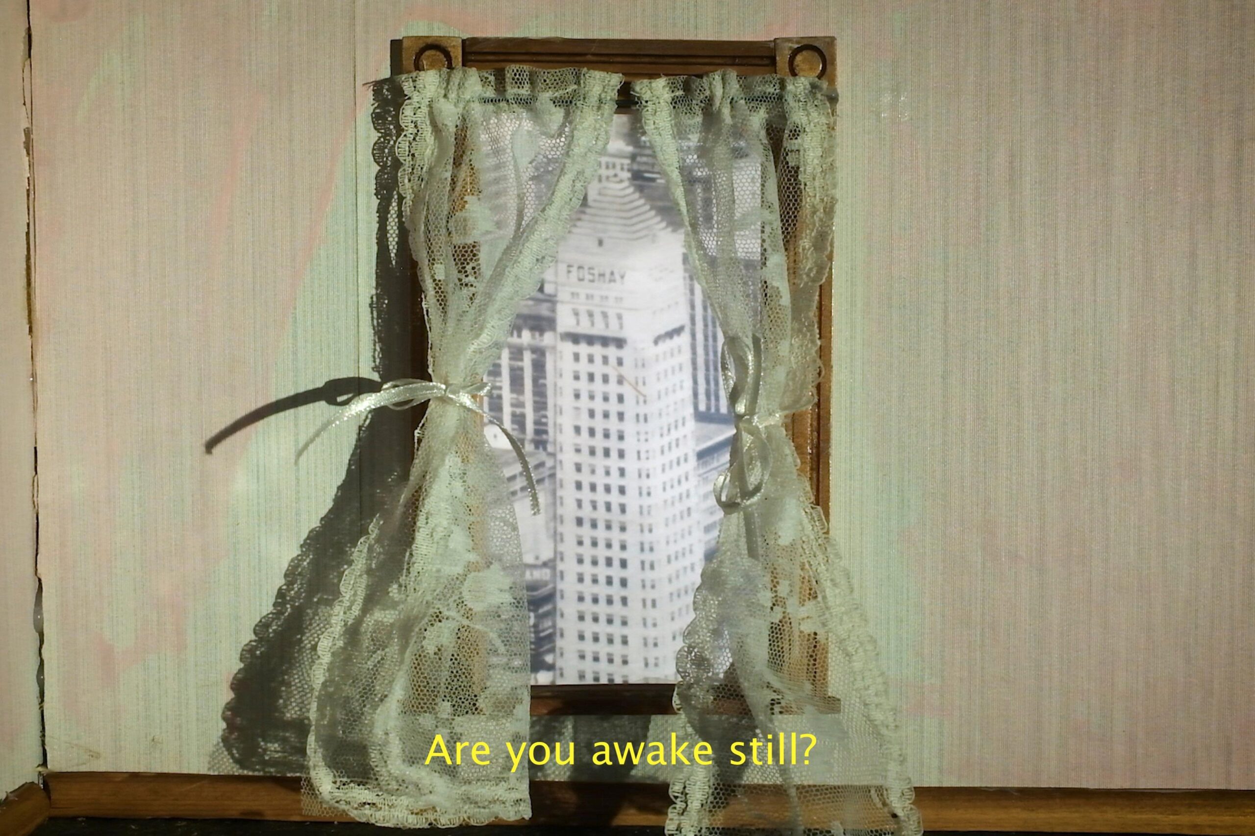 The image shows a window with intricate lace curtains tied back on either side. Through the window, a black-and-white image of a tall, white building with the word 'FOSHAY' visible on its facade is seen. Below the window, there is text in yellow that reads, 'Are you awake still?'