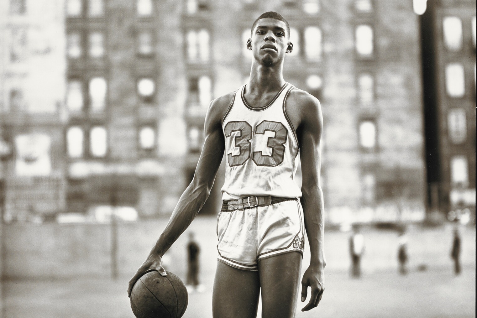 A black-and-white photo of a young basketball player standing on an outdoor court. The player is wearing a sleeveless basketball jersey with the number 33 on the front and matching shorts. He holds a basketball in one hand by his side. The background reveals blurred buildings.