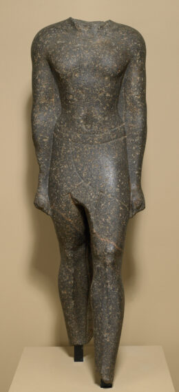 Stone statue depicting a human figure without a head. The figure stands upright, showcasing detailed anatomical features of the torso, arms, and legs. The material of the statue has a mottled texture with dark and light spots.