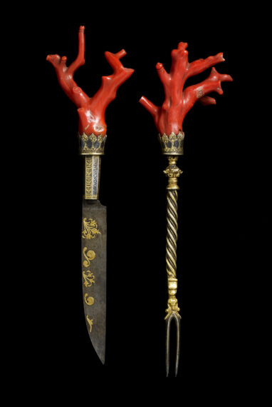 Ornate knife and fork with red coral handles on black background.