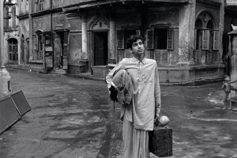 Black-and-white image depicting a person standing on a deserted street in front of a worn, multi-story brick building with shuttered windows. The person is wearing traditional attire, consisting of a long kurta and loose pants, and is holding a folded piece of clothing and a suitcase.