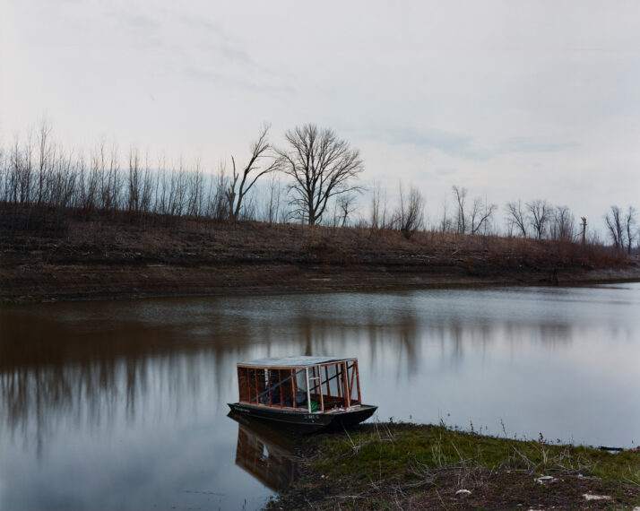 Photo of a small wooden boat run up on the shore of a calm river surrounded by a barren shoreline.