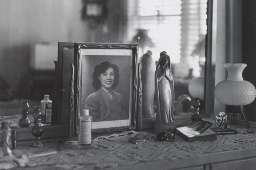 Black and white photo of a dresser with a portrait, bottles, and figurine