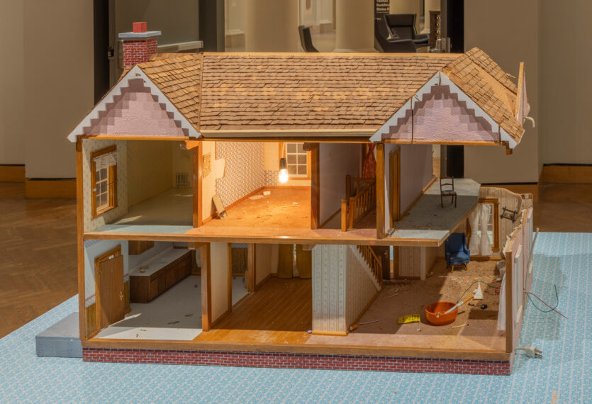 The image shows a detailed dollhouse with its front wall removed, revealing the interior of a two-story structure. The house has a shingled roof and brick-like foundation. 
