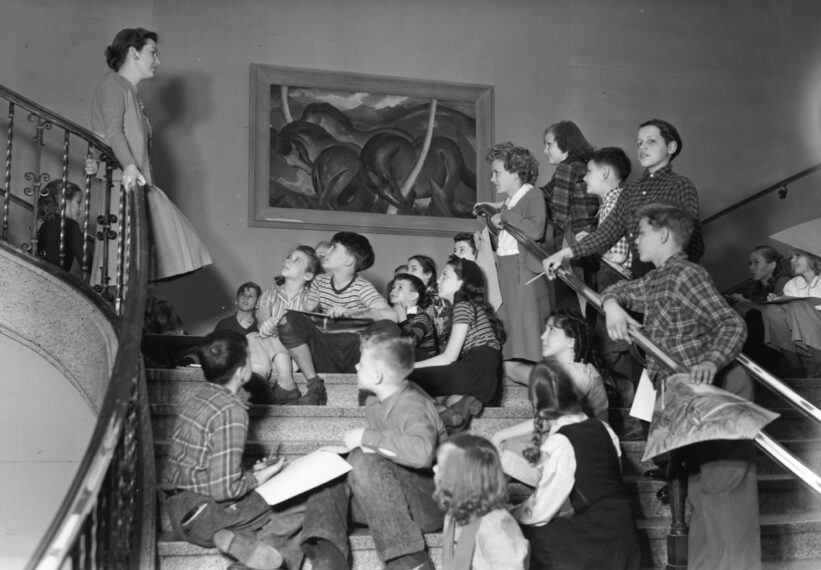 A black-and-white photo of a group of children sitting on a staircase, looking up at a woman standing at the top of the stairs where a large framed painting hangs on the wall.