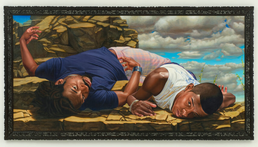 Two men lying closely on a rocky surface beneath a sky filled with dramatic clouds. The man on the left, wearing a dark blue shirt, lies with his head tilted and one arm bent upward. His hair is styled and he has facial hair. The man on the right is dressed in a white sleeveless shirt. Both men are lying on their sides and their bodies are slightly intertwined.