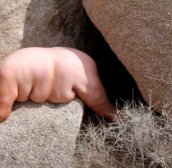 The image shows a naked Caucasian person attempting to squeeze through a narrow gap between large, rough-textured rocks. The person's back is towards the viewer, and their legs are extended into the dark crevice.