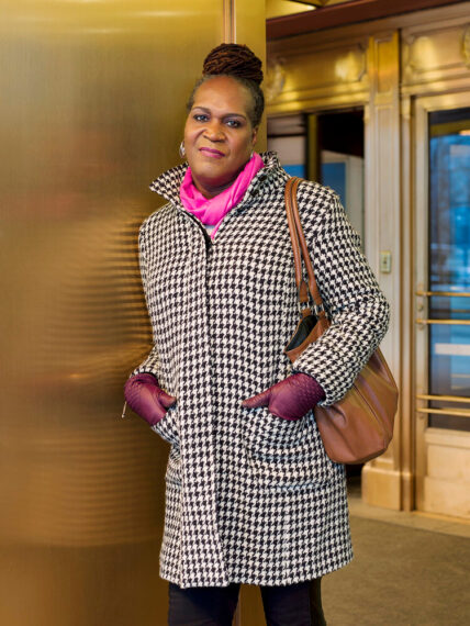 Woman in houndstooth coat and pink scarf leaning against by a gold wall.