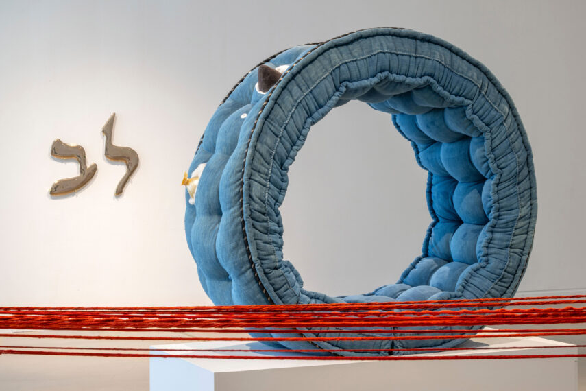 Blue circular fabric sculpture next to golden Hebrew letters on wall.