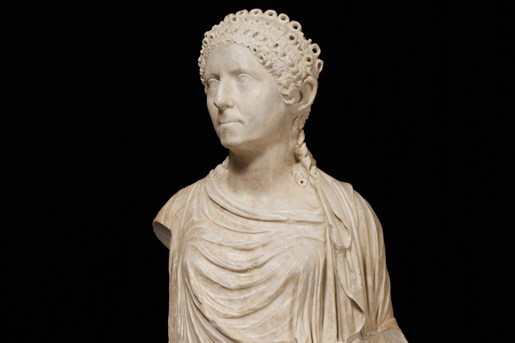 Marble statue of a draped figure with curly hair set against a black background.