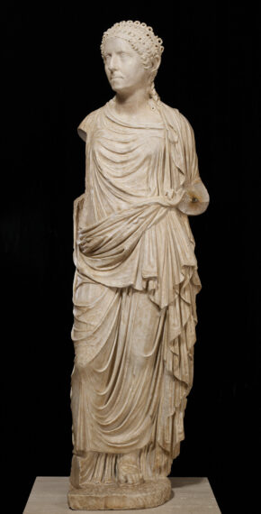 Marble statue of a draped figure with curly hair set against a black background.