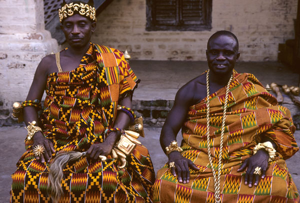 Two men wearing colorful, patterned robes and gold jewelry sit outside.