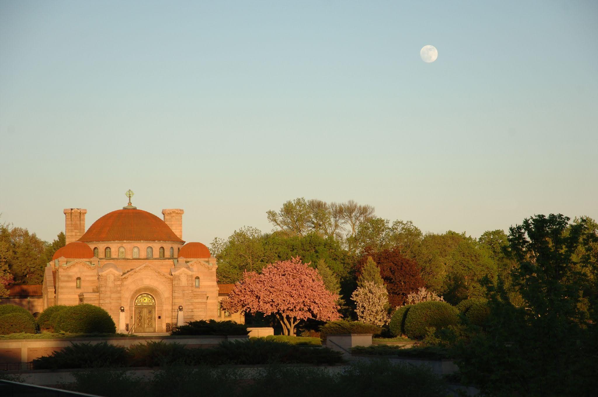 Building with red dome and moon in a clear sky above trees.