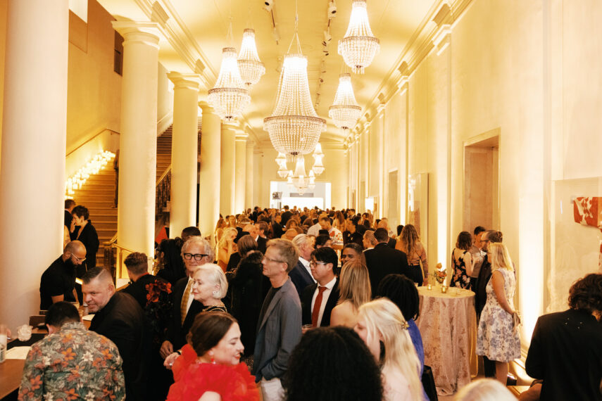 The image shows a crowded formal event taking place in an elegant hall. The hall features high ceilings with several large, ornate chandeliers. There are many people dressed in formal attire engaged in conversations.