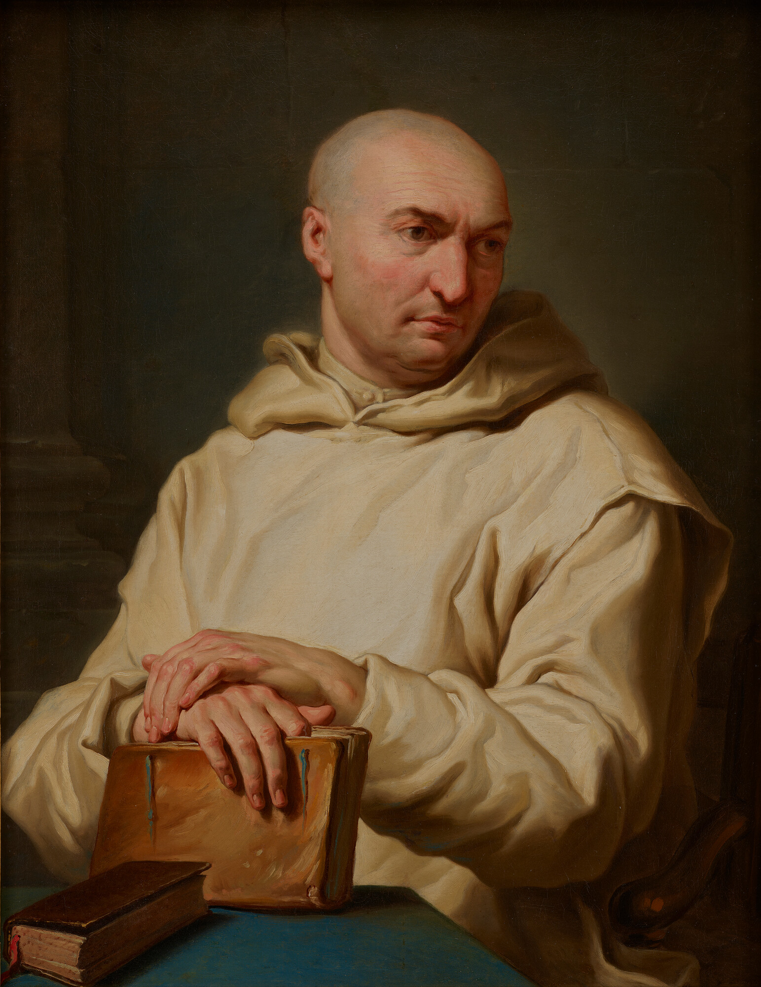 The image depicts a bald person wearing a light-colored robe. They are sitting and have their hands resting on the top of a closed leather book on a table.