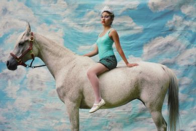 young woman wearing a turquoise tank top, turquoise and black skirt, white shoes and white beaded headdress, seated bareback on a white speckled horse, standing in front of a backdrop painted with blue sky and white clouds