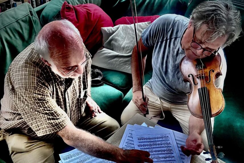 Two people are sitting together on a green couch, focusing on sheet music spread out on a table before them. The person on the left is pointing to the sheet music. The person on the right holds a violin.