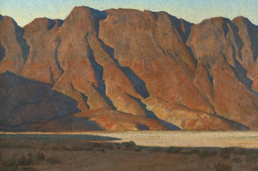 The image shows a landscape featuring a rugged mountain range with reddish-brown hues under a light blue sky.