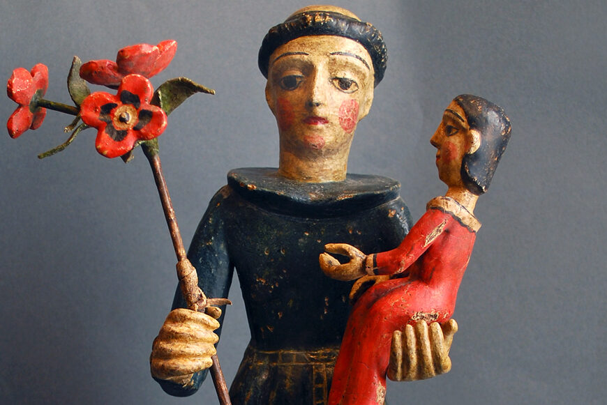 The image shows a carved wooden figure with painted features, wearing dark clothing. The figure holds a red flower on a long stem in one hand. In the other hand, it cradles a smaller figure, painted red, resembling a child or doll.