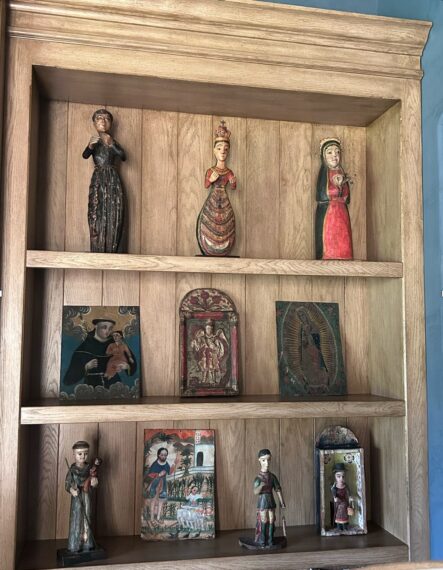 The image shows a wooden shelf against a wall, divided into three sections. The top shelf has three wooden statues. The middle shelf contains three paintings. The bottom shelf has two small wooden statues and two small paintings.
