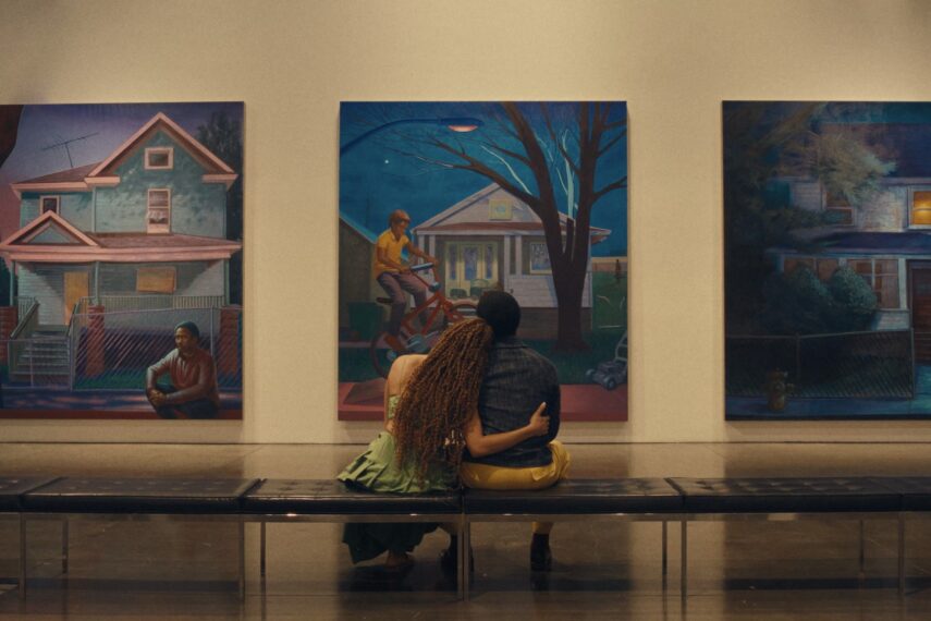Photo of two people sitting closely together on a bench in a gallery, facing a wall with three large paintings.