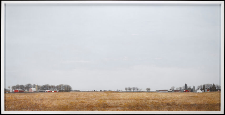 The image features a large, open field with dry grass stretching across the foreground. In the distance, there is a line of farm buildings, including several red and white barns and a few silos. Trees with bare branches dot the horizon.
