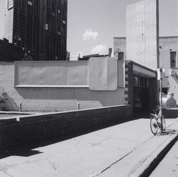 The black-and-white photo shows an urban setting with tall buildings in the background and a smaller, one-story structure in the foreground.