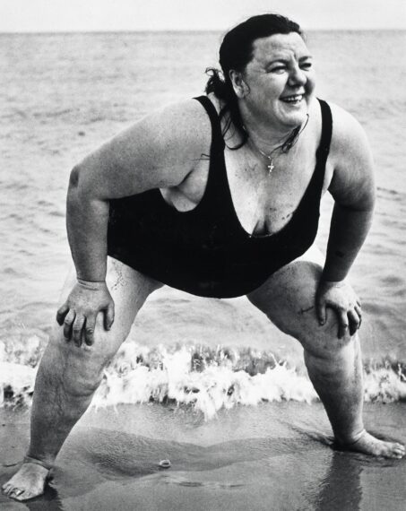 A black-and-white photograph shows a person standing at the shore in a black swimsuit. They are leaning forward with hands resting on their knees and are smiling widely. The ocean waves lap against their legs, and the horizon stretches in the background.