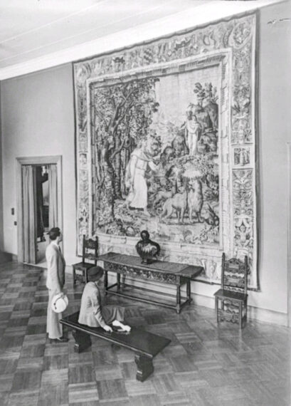 Two people view a large tapestry on a gallery wall in a museum.
