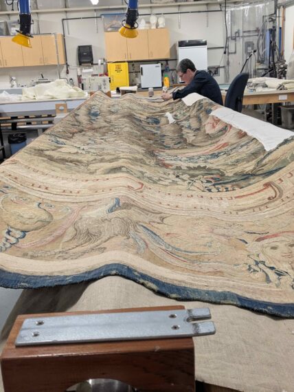 In a workshop setting, a person sits at a table, restoring a large, ornate tapestry that spreads across the entire tabletop.