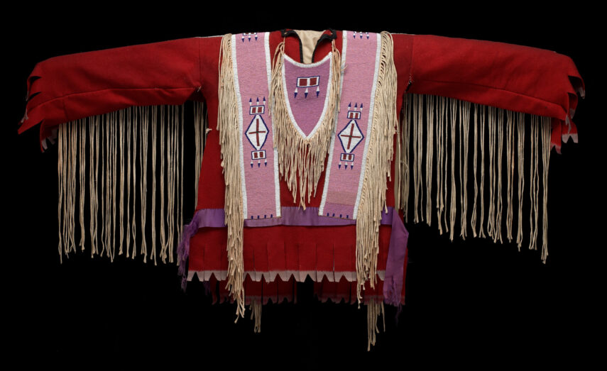 Image shows a traditional garment against a black background. The garment is predominantly red with long sleeves. It features beige fringe hanging from both the sleeves and the front. The center has two wide vertical pink panels with white and purple geometric patterns, including diamonds and rectangles.