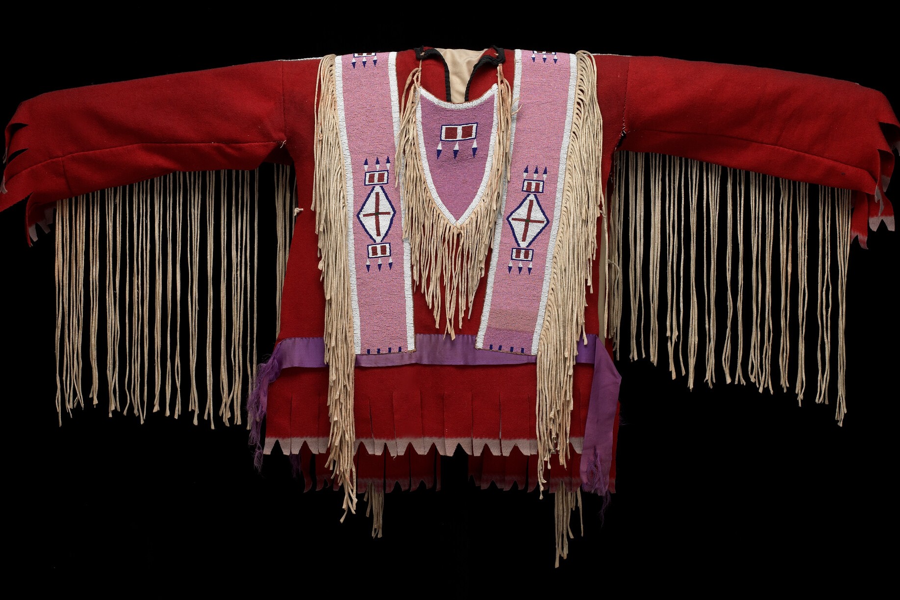 Image shows a traditional garment against a black background. The garment is predominantly red with long sleeves. It features beige fringe hanging from both the sleeves and the front. The center has two wide vertical pink panels with white and purple geometric patterns, including diamonds and rectangles.