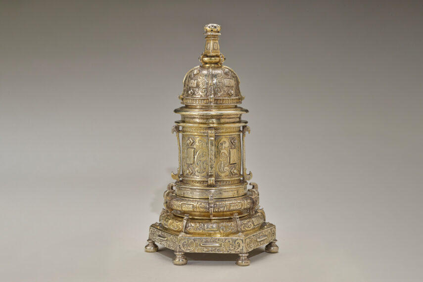 The image shows an intricate golden object with a domed top with detailed patterns, supported by cylindrical sections featuring engravings and decorative motifs. The base is square with four small, rounded feet, also adorned with intricate designs.