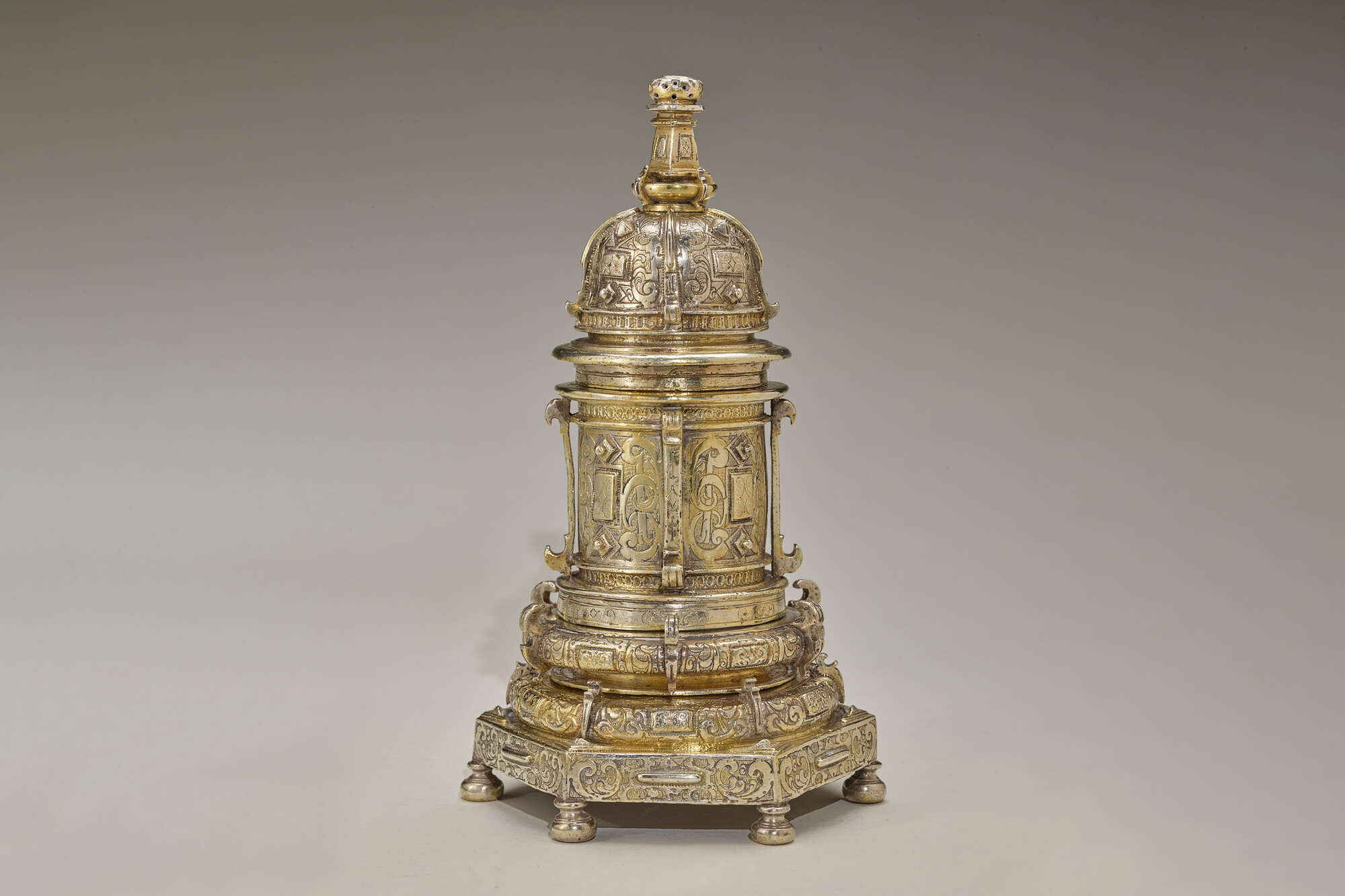 The image shows an intricate golden object with a domed top with detailed patterns, supported by cylindrical sections featuring engravings and decorative motifs. The base is square with four small, rounded feet, also adorned with intricate designs.