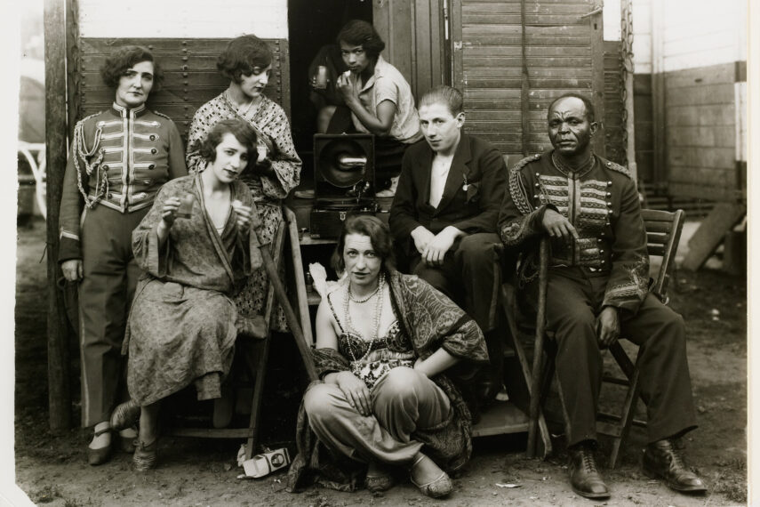 The black-and-white photograph shows a group of seven people posing in front of a wooden door. Some individuals are sitting while others stand.