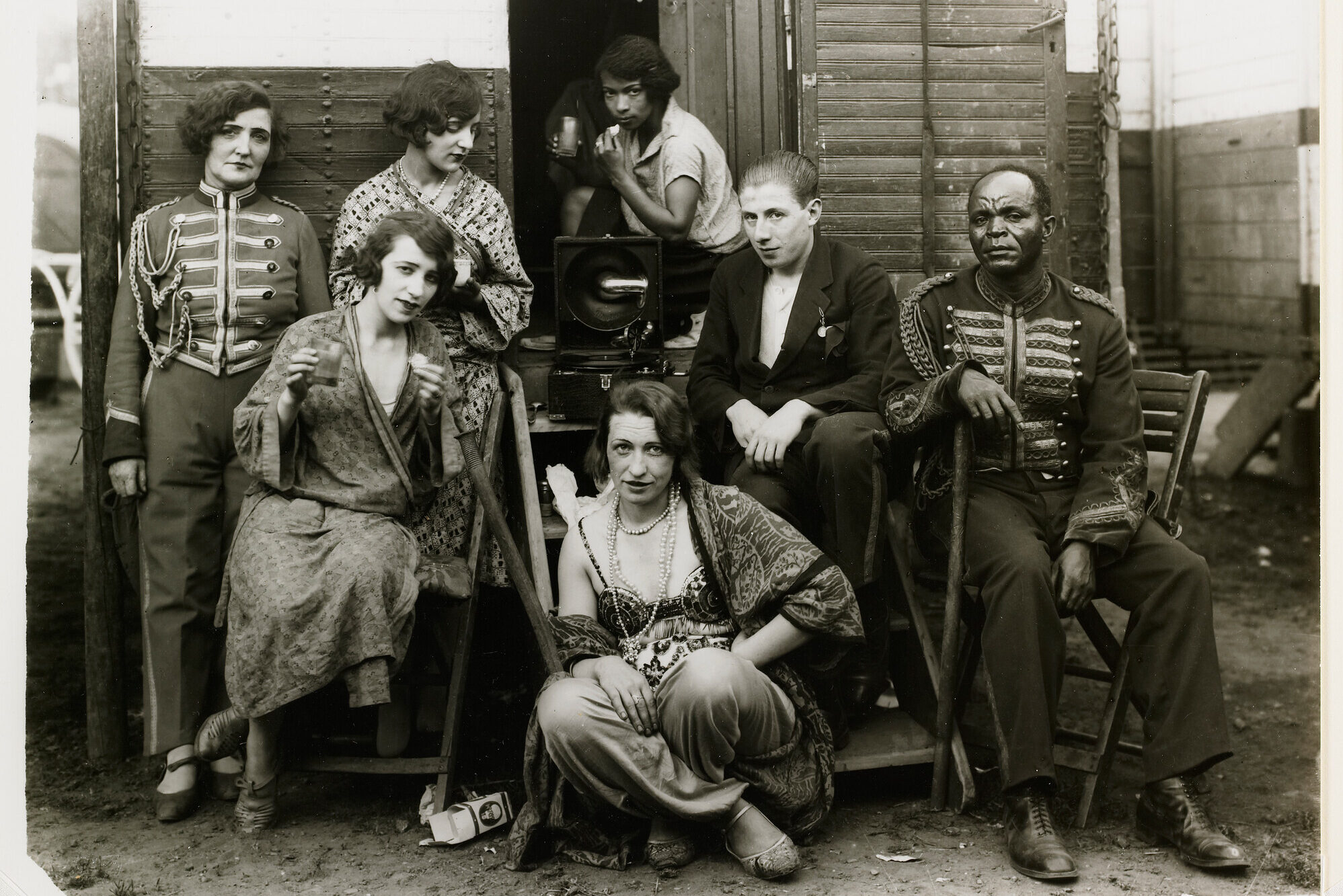 The black-and-white photograph shows a group of seven people posing in front of a wooden door. Some individuals are sitting while others stand.