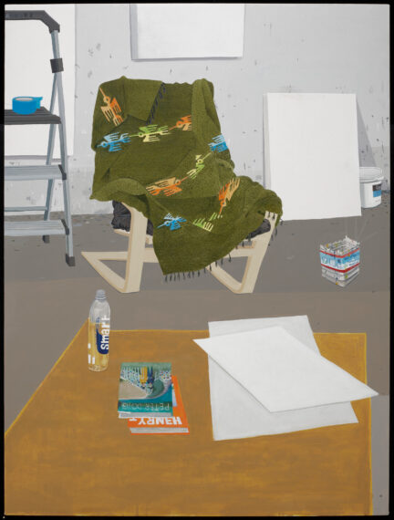 The image shows a room with a light beige folding chair covered by a green blanket with colorful abstract patterns. To the left, a silver step ladder holds a blue bowl on its top shelf. In the background, a white canvas is propped against a wall. On the right side, a container of paint sits on the floor. In the foreground, a wooden table has two books, some papers, and a clear plastic water bottle on top. 