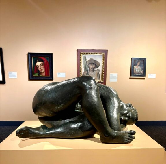 Bronze sculpture of a crouched figure in an art gallery with paintings on the wall.