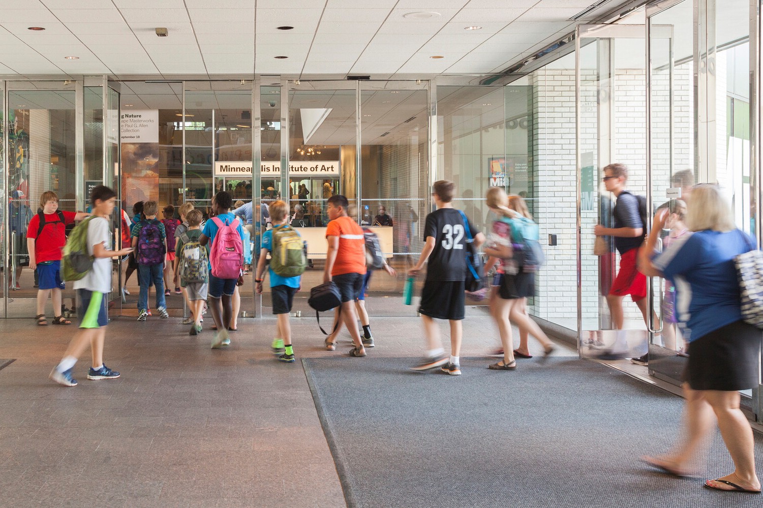 Photo of a large group of people, mainly children, walking through a set of large glass doors into a building. Most are wearing casual summer clothing and backpacks.