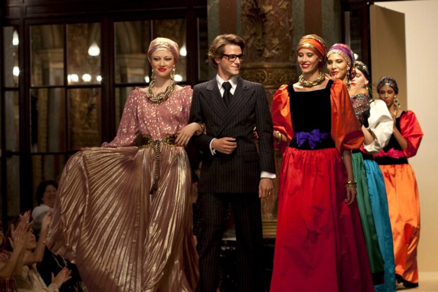 The image shows a fashion show scene where a male figure, dressed in a dark pinstripe suit and glasses, walks down the runway linked arm-in-arm with two female models wearing elaborate gowns.