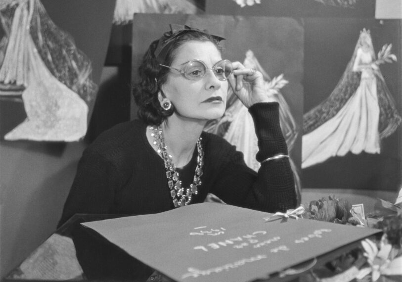 Black-and-white photo of Coco Chanel wearing glasses and jewelry, surrounded by drawings of bridal gowns.