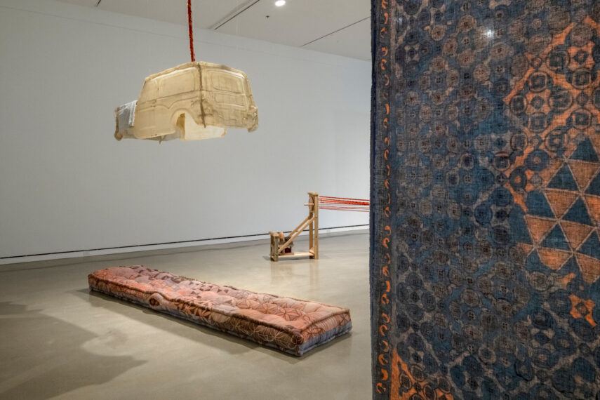 Art installation with hanging car, long cushion, loom, and patterned textile.