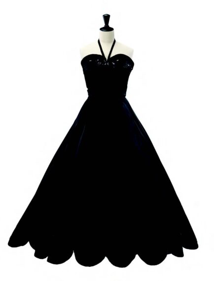 A black gown displayed on a mannequin, with a fitted bodice with a sweetheart neckline and thin straps creating a halter neck style. The skirt is full and flares out from the waist, featuring a scalloped hemline.