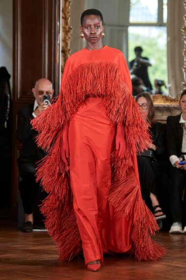The image shows a model walking down a runway in a long red dress with extensive fringe detailing.