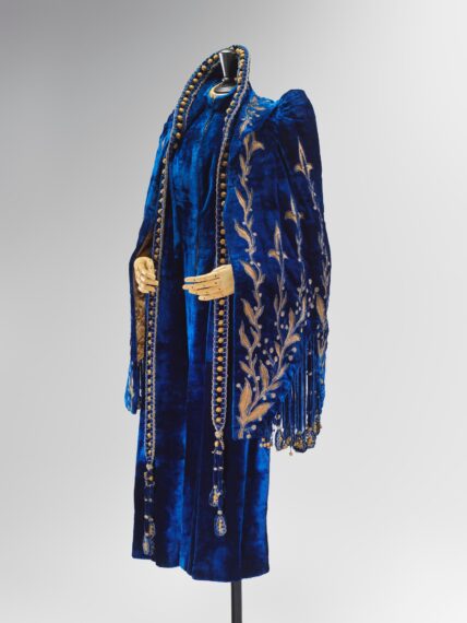 A long, royal blue velvet coat displayed on a mannequin. It features extensive gold embroidery in a leaf motif, beaded trim along the edges, and tassels.